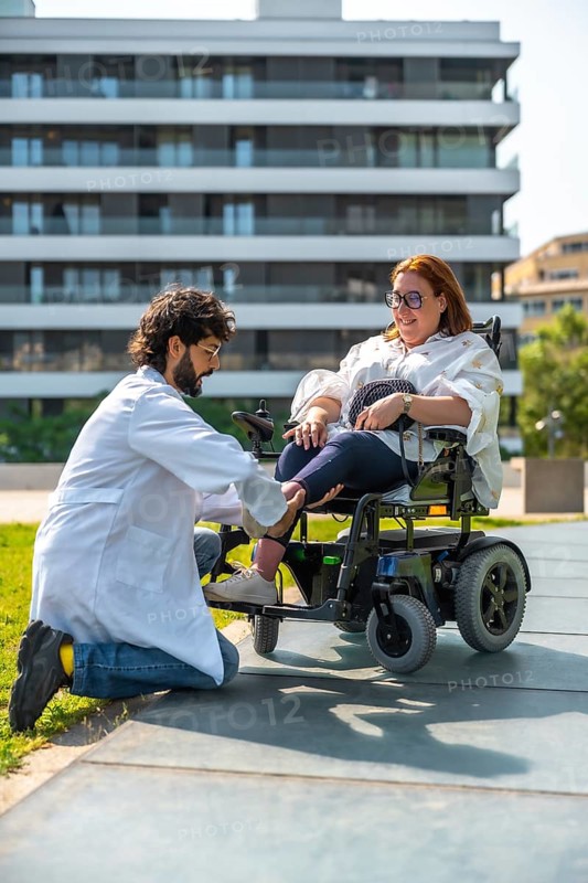Doctor adjusting footrest of electric wheelchair for smiling disabled woman in park, promoting accessibility and healthcare