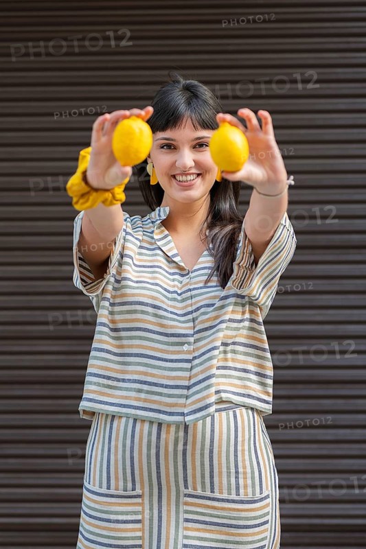 A cheerful woman in a striped outfit holds two bright lemons in front of her. Her vibrant smile and colorful attire create a lively and joyful atmosphere in this outdoor setting