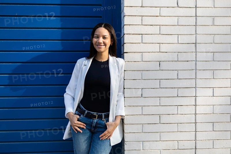 A confident woman with a casual business look, wearing a white blazer and jeans, stands against a blue and white wall backdrop, embracing a modern lifestyle