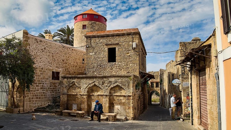 Older buildings in an old town with a red tower and people on benches, Rhodes Old Town, Rhodes, Dodecanese, Greek Islands, Greece