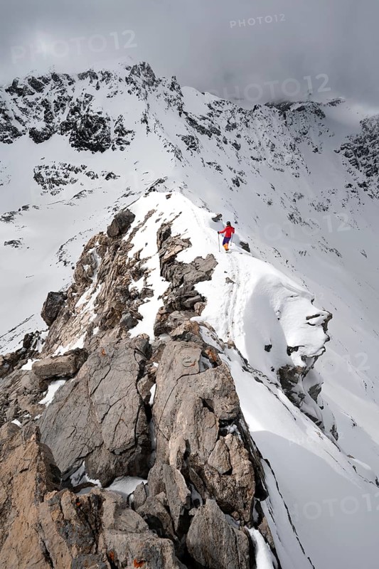 Mountaineer descending the narrow ridge of Piz Laviner, view of mountain panorama in wintry mountain landscape with dramatic clouds, Grisons Haute Route, Albula Alps, Rhaetian Alps, Grisons, Eastern Switzerland, Switzerland