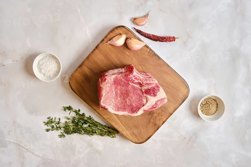 Raw pork loin rack joint on a wooden cutting board. Garlic cloves, dried chili, and fresh thyme are arranged around it. The scene is set on a marble countertop, ready for meal preparation