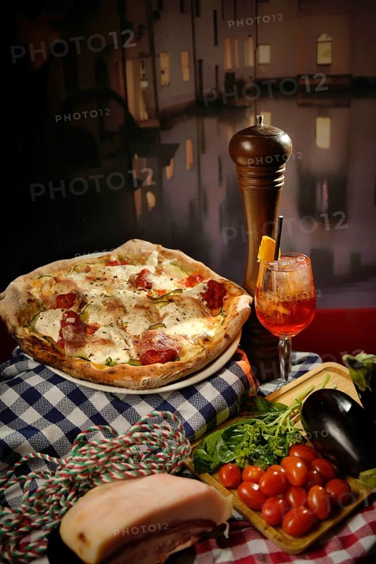 A delicious bustling Italian pizza adorned with mozzarella and ripe cherry tomatoes, laid out beside fresh ingredients like basil, arugula, and eggplant, with a rustic pepper grinder and a glass of spritz in the background, emphasizing a cozy dinnertime setup