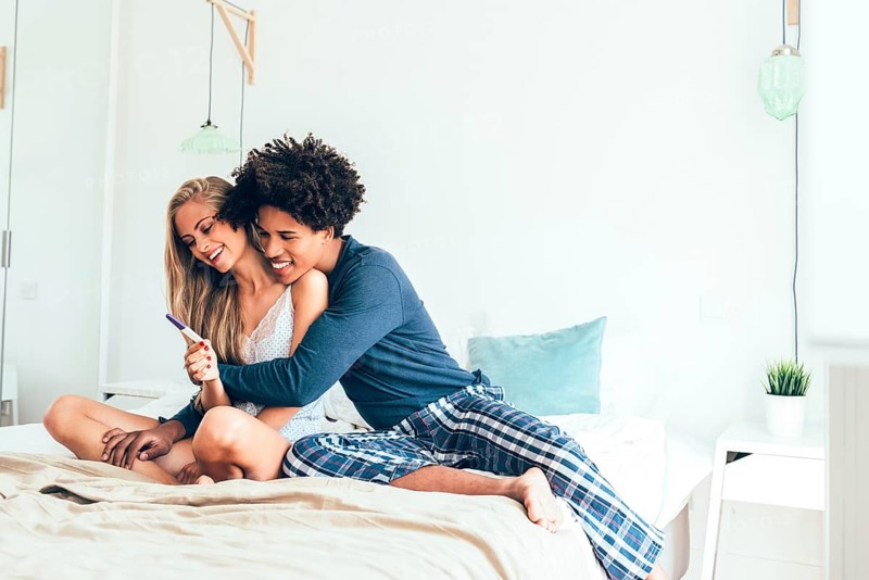 A happy couple sits on their bed, hugging and smiling as they celebrate a positive pregnancy test result. The scene radiates excitement and love in a cozy, modern bedroom