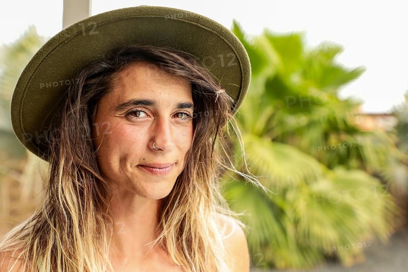 Close-up portrait of a woman with a hat and nose ring, smiling gently outdoors Vibrant greenery in the background complements her natural and relaxed expression
