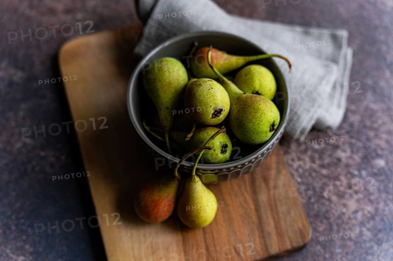 A rustic arrangement featuring a bowl filled with fresh wild pears. The pears exhibit a mix of green and red hues, resting on a wooden board with a cloth napkin