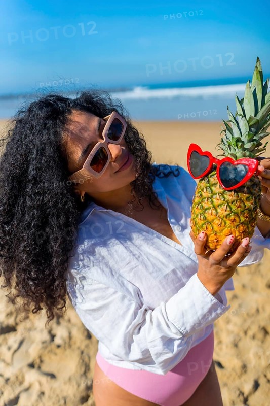 Woman with curly hair and sunglasses standing on a sunny beach, holding a ripe pineapple adorned with heart shaped red sunglasses, embodying summer vacation and tropical fun