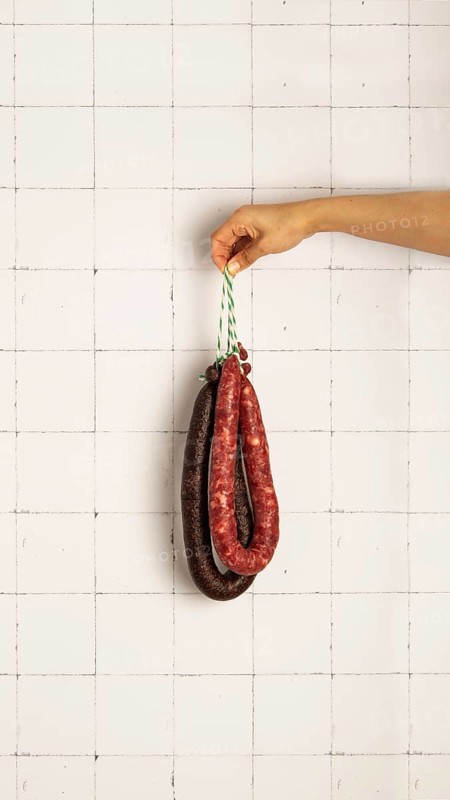 A hand holds black pudding and pork sausage against a white tiled wall, showcasing different textures and colors. The composition highlights culinary tradition and artisanal meat products