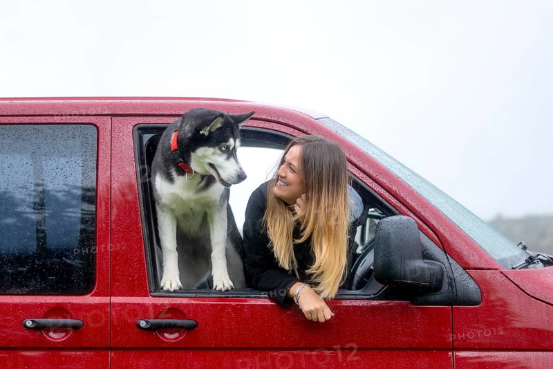 A woman and her dog share a moment through the window of a stationary red van on a rainy day, displaying a strong bond of friendship and adventure