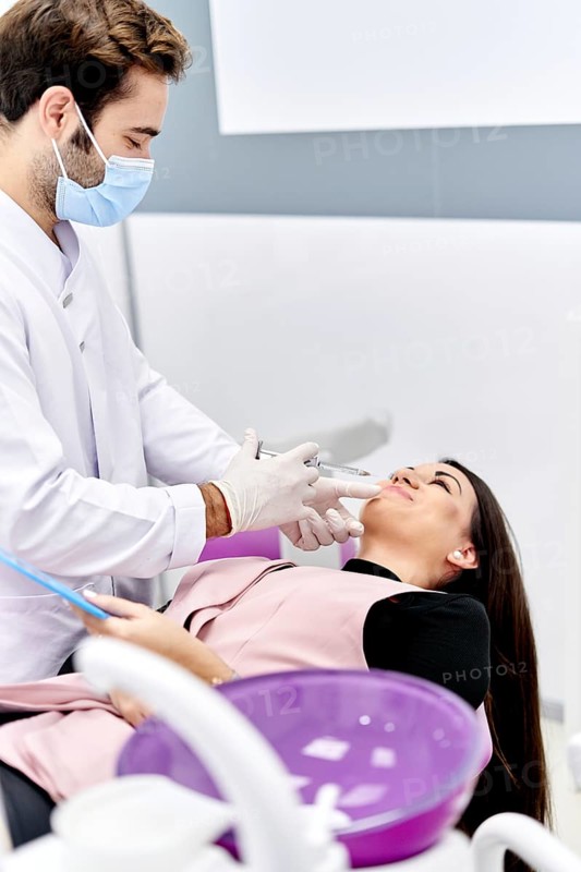A beautician, wearing a face mask and gloves, gently injects hyaluronic acid into a patient's face to enhance skin appearance in a clinical setting, focusing on precision and care