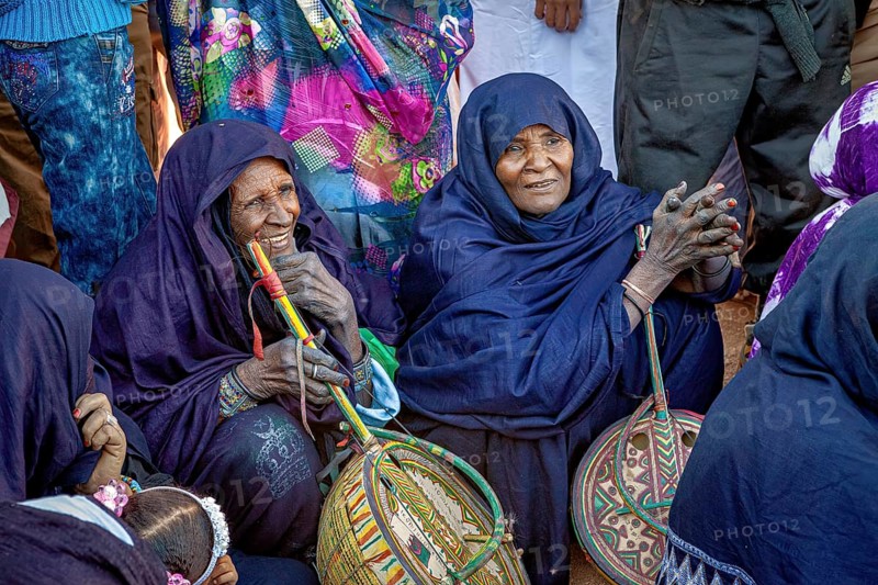 Women in traditional dress sit together holding decorated baskets, surrounded by a group of people, Traditional woman play music at the Tuareg Festival of Tamanrasset in the Sahara in Algeria