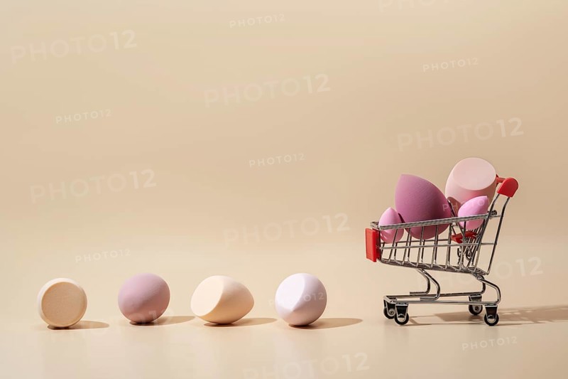 Soft cosmetic sponges in various pastel tones are displayed creatively in a miniature shopping cart against a neutral background, highlighting their texture and versatility
