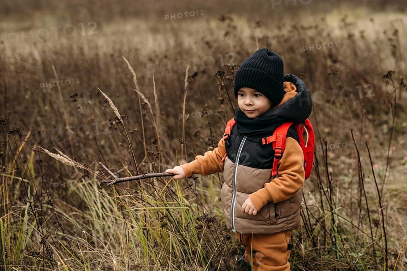 Young boy wearing a warm jacket and backpack explores a field during an autumn walk. He holds a stick, surrounded by tall grasses, enjoying the crisp fall air and scenery