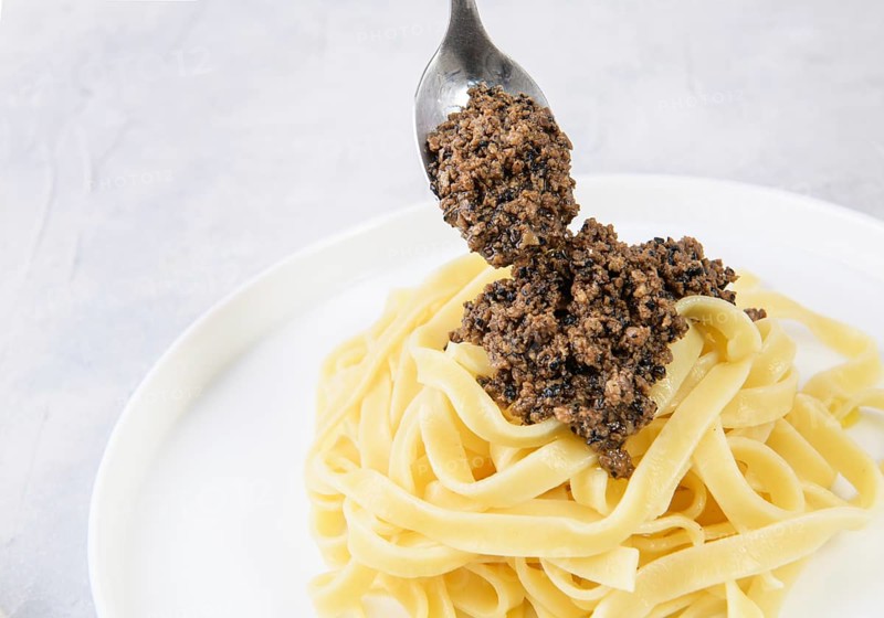 A spoon of savory black truffle sauce being added to a bowl of fresh pasta. Perfect for a gourmet culinary experience, capturing the essence of Italian cuisine