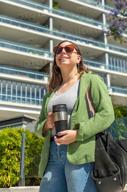 A young woman wearing sunglasses and casual clothes, holding a travel mug, smiling outdoors near a modern residential building with greenery, captured on a bright sunny day