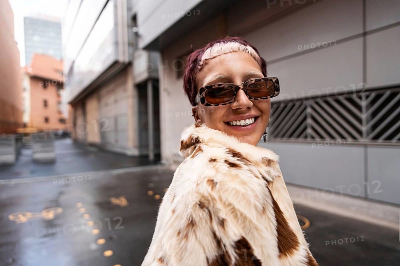 A stylish woman wearing sunglasses and a fur coat smiles confidently in an urban setting. Her chic hairstyle and confident aura add to the fashion-forward atmosphere