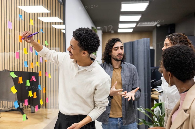 A diverse group of coworkers engages in a brainstorming session, using vibrant sticky notes on a glass wall in a modern office. Collaboration and creativity are emphasized