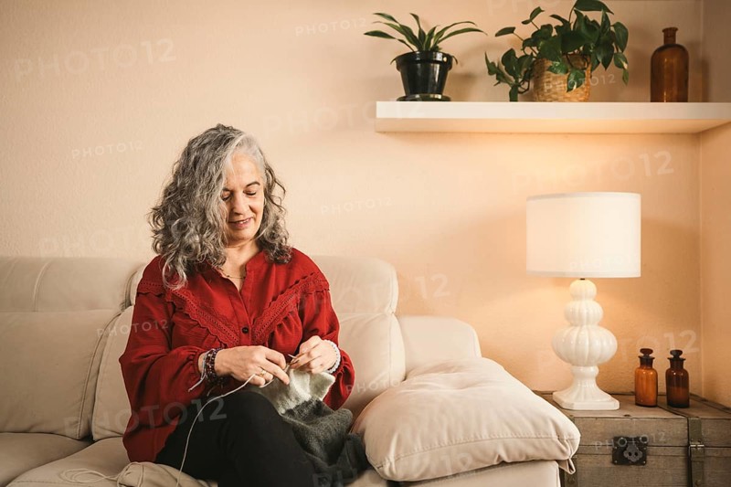 A woman with gray hair sits comfortably on a sofa, knitting with focus. The cozy living space features plants and a lamp, creating a warm and inviting atmosphere
