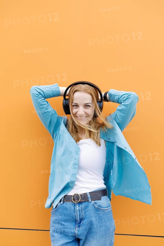 A woman in casual attire, smiling with headphones on, stands against a bright orange background in Warsaw, effortlessly combining style with technology