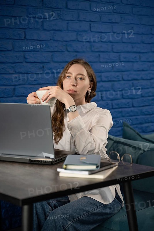 A woman sits at a table with a laptop, enjoying a tea break. She wears a white blouse and has books and glasses on the table, set against a blue brick wall