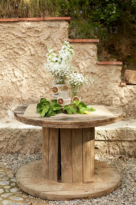 A rustic wooden table features an elegant floral arrangement with white flowers in glass jars. Accented with greenery and dried citrus, itâ€™s perfect for a boho chic wedding decor