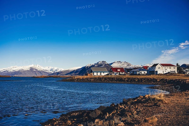 Picturesque coastal village in Stokksnes, Iceland, with brightly colored houses along a rocky shoreline, framed by distant snow-capped peaks and a clear blue sky