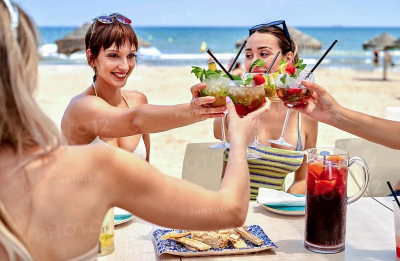 Friends gather at a beach side table, raising colorful cocktails under the summer sun. The ocean sparkles in the background, creating a joyful atmosphere for their day