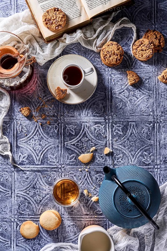 A comforting arrangement featuring a steaming cup of coffee, a pot of tea, chocolate chip cookies, butter cookies, and an open book, all on a patterned blue tablecloth