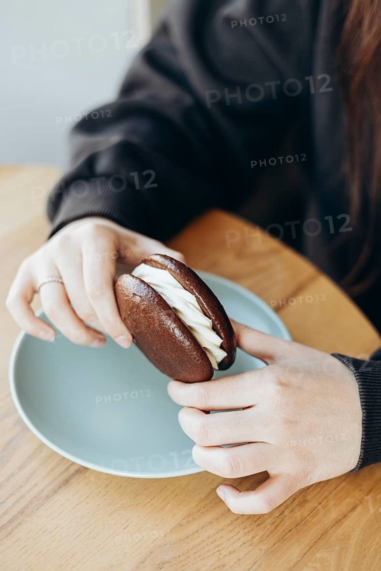 A close-up of hands holding a chocolate whoopie pie with creamy filling atop a light-colored ceramic plate on a wooden table, capturing a sweet treat moment