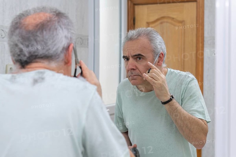 A man carefully trims his hair in front of a bathroom mirror. He holds scissors, focusing on achieving a neat appearance. The ambiance is casual and personal grooming