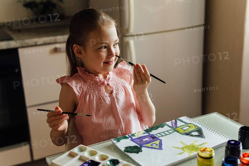 A young girl in a pink dress happily paints a picture with watercolors on paper at a kitchen table. Sunlight gently illuminates her focused expression and creative process