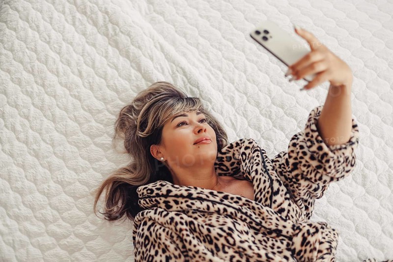 A woman in a cozy leopard print robe lies comfortably on a bed with a white quilt, using her smartphone. She appears relaxed and engaged with her device