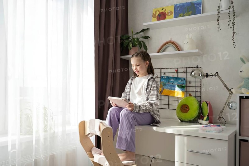 A young girl sits barefoot on a white desk in her sunlit bedroom, focused on using a digital tablet. Surrounded by soft colors, plants, and playful decor, the scene feels creative and calm