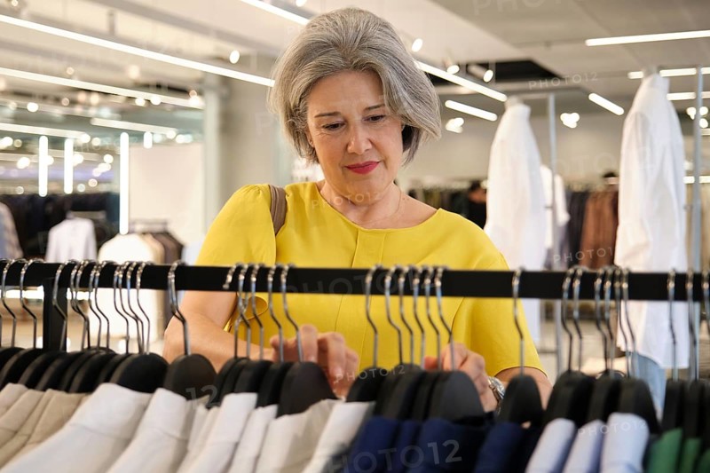 Mature woman with gray hair shopping in a modern clothing store. She wears a yellow blouse and browses a rack of clothes, focused on her selection in a bright setting