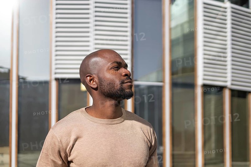 A cuban man in a beige sweater stands confidently outside a modern building with a backdrop of glass and metal. Sunlight highlights his thoughtful expression and strong features