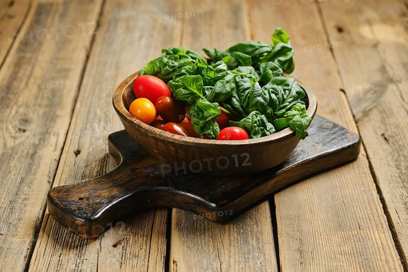 A wooden bowl with red and yellow cherry tomatoes along with fresh green basil leaves. The bowl is placed on a wooden cutting board. This scene takes place in a rustic kitchen setting