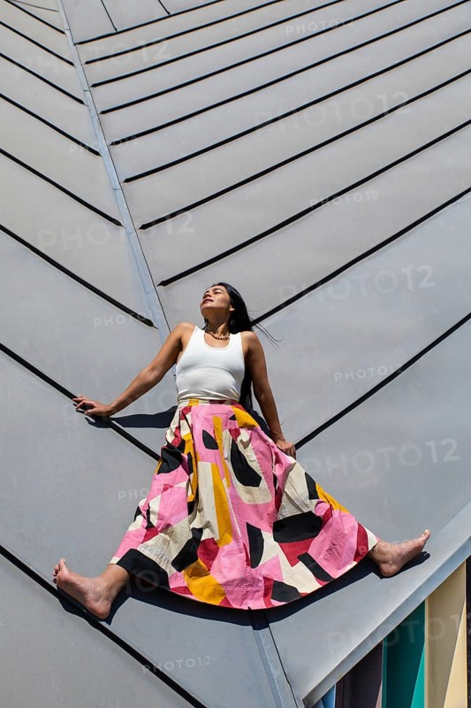 A woman in a vibrant, colorful outfit poses against the backdrop of modern architecture. The bright clothing contrasts with the sleek lines of the urban environment