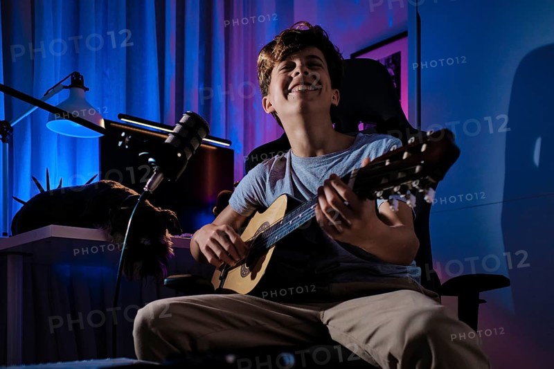 A young musician joyfully strums a guitar in a creatively lit room, surrounded by a microphone and vibrant ambient lighting, capturing the essence of music and creativity