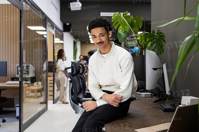 A modern office setting with a man casually sitting and colleagues conversing in the background. Greenery and sleek technology enhance the contemporary workspace