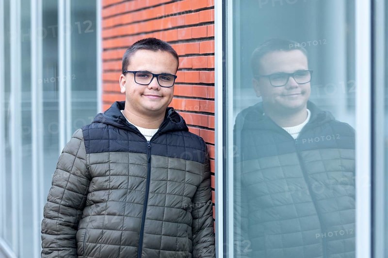 A young man, wearing glasses and a casual jacket, leans against a window at a mental health leisure center. He appears relaxed and contemplative, reflecting the themes of mental health awareness