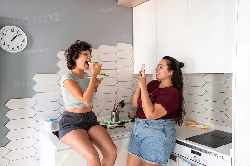A warm scene of a lesbian couple in a kitchen, one taking a photo while the other enjoys a bite of avocado toast, capturing a cozy morning routine