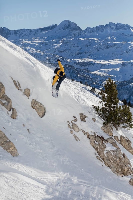 A snowboarder in a dynamic pose navigates a steep, snowy slope against the majestic backdrop of rugged mountains. The scene captures the thrill and beauty of winter sports