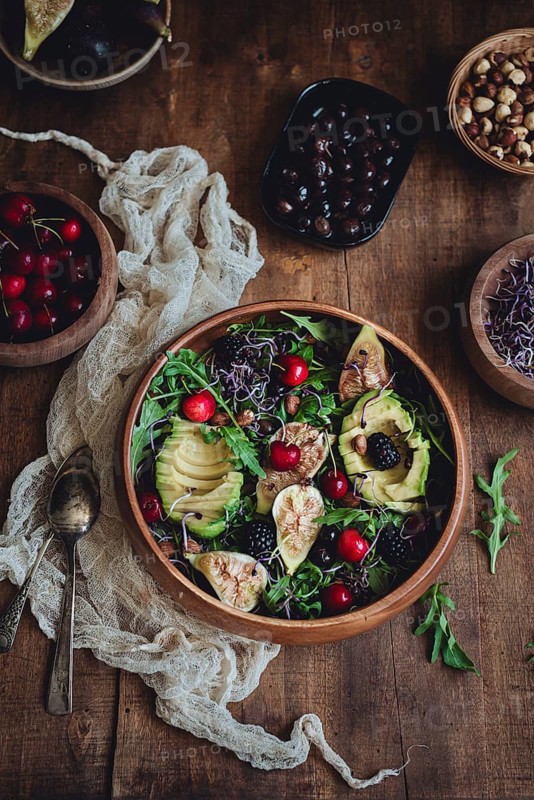 A vibrant salad featuring figs, avocado, cherries, arugula, and blackberries in a wooden bowl. The scene is complemented by various nuts and fruits surrounding the main dish