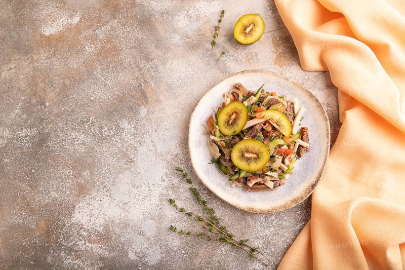 Meat salad with beef kiwi and vegetables on brown concrete background and orange linen textile. top view, flat lay, copy space