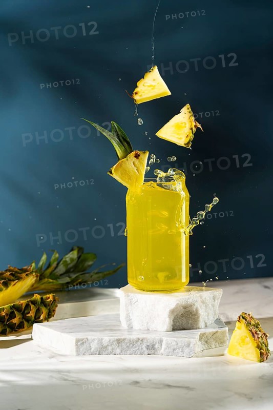 A vibrant pineapple drink in a glass jar with slices of pineapple splashing over it on a marbled platform against a dark blue background