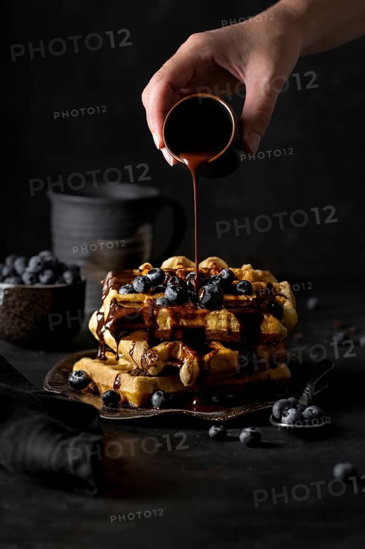 Crop anonymous person adding chocolate syrup from small cup on top of delicious waffles pyramid with blueberries on kitchen