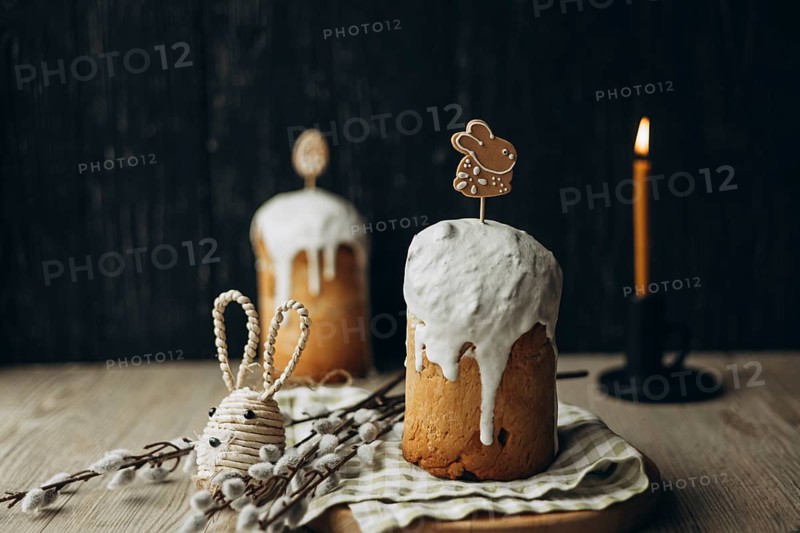 Traditional Russian Easter kulich, a tall bread topped with frosting, decorated with a festive gingerbread bunny and accompanied by pussy willow catkin branches, an Easter bunny figure, and a lit candle against a dark wooden background