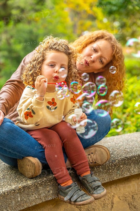 Vertical photo of a curious girl blowing bubbles in the park sitting with her mother