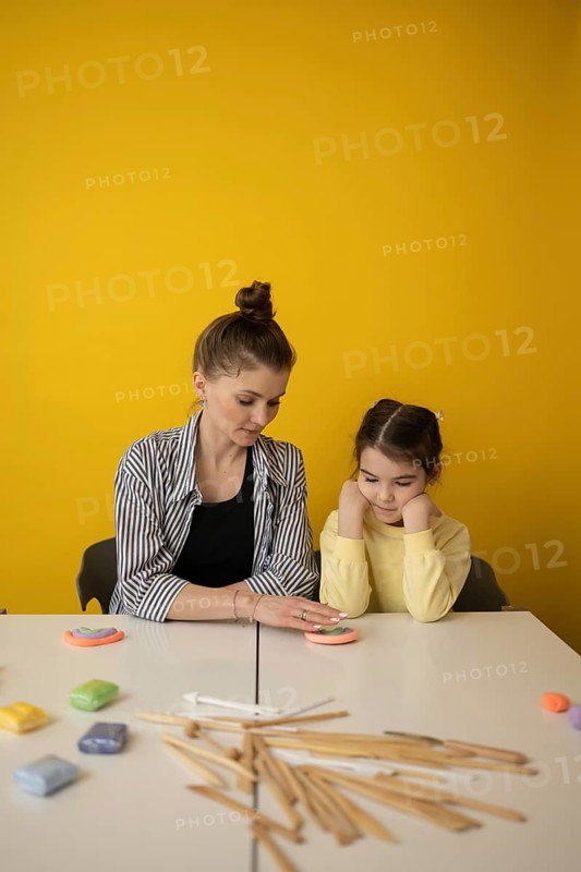 A teacher and child engage in a creative art lesson with colorful materials on a white table against a vibrant yellow background, fostering imagination and learning