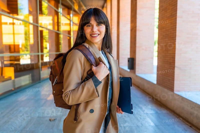 Portrait of a young woman carrying a backpack and tablet, walking through a modern university corridor, representing education, learning, and student life